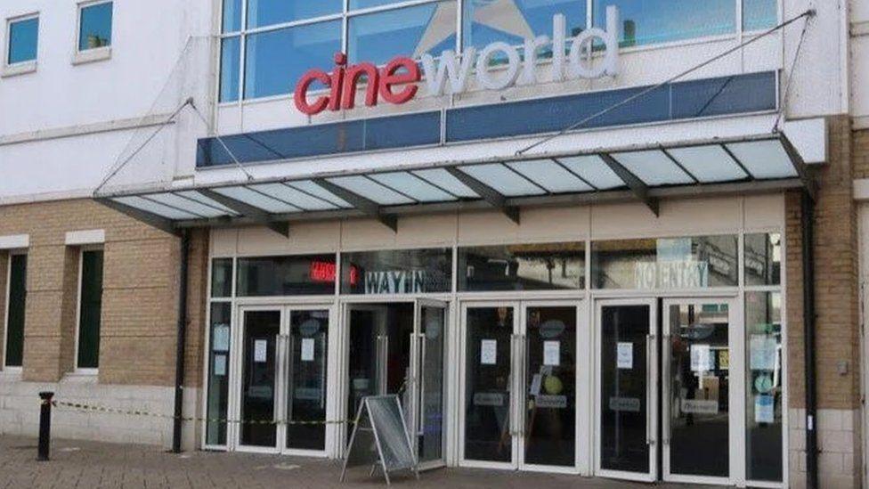 Entrance to cinema with multiple doors and A frame sign outside a paved street.