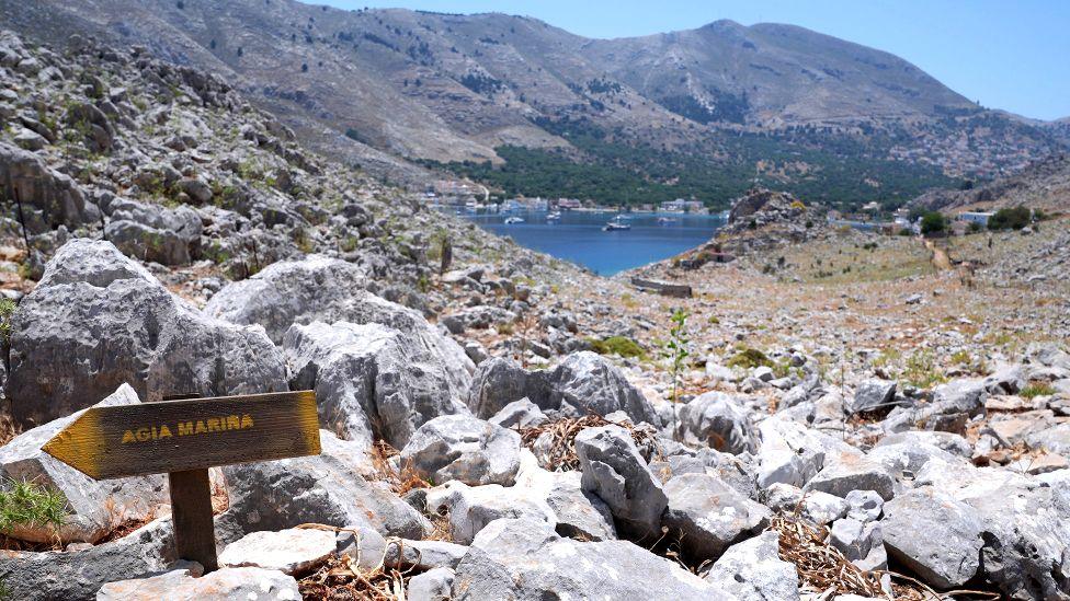 A direction sign on a rocky path in the hills of Pedi (Pedi centre pictured in the distance, right), a small fishing village in Symi, Greece, pointing toward Agia Marina on 8 June