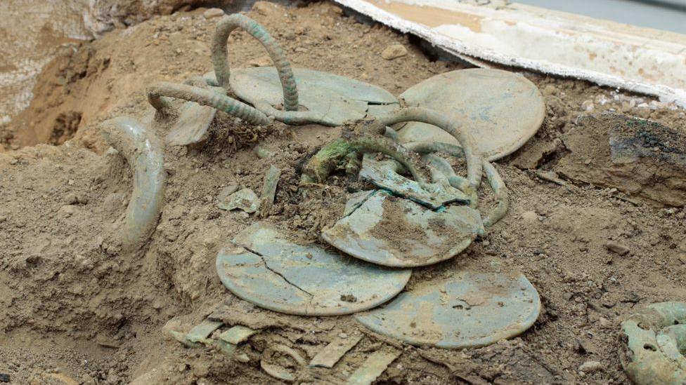 Bronze Age artefacts jut out of the dirt with a range of circular items and hoops sticking out