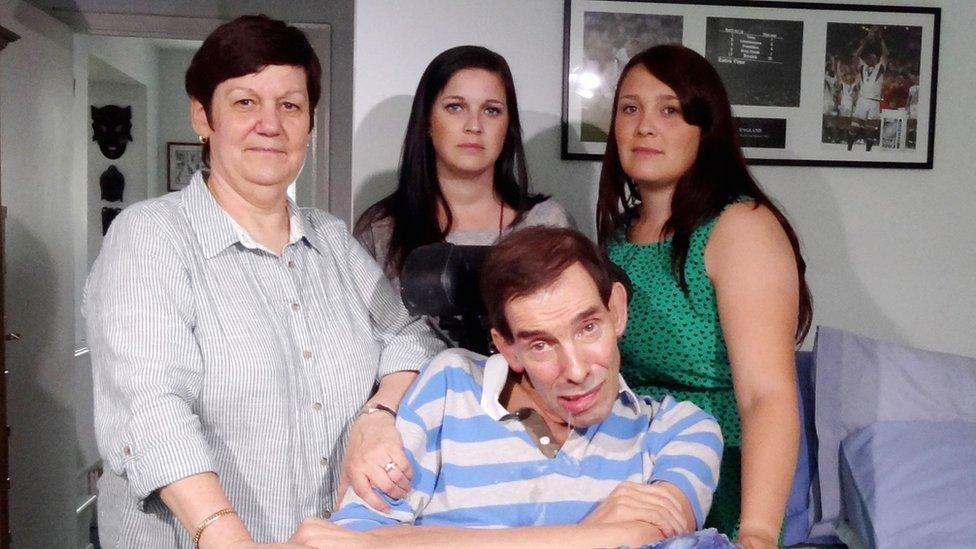 Tony Nicklinson sitting in his living room, wearing a white and blue striped shirt with his arms crossed. He is surrounded by three of his family members, who are looking at the camera with a solemn expression. 