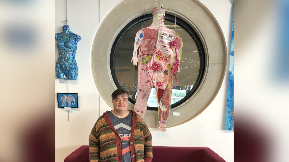 Allie Watson smiling at the camera while standing in front of her textile creation, which is a human body.