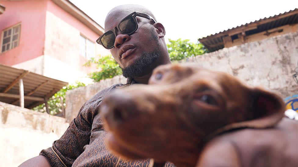 Iyke Elueze wearing sunglasses hugs his brown dog whilst sitting and looking at the camera
