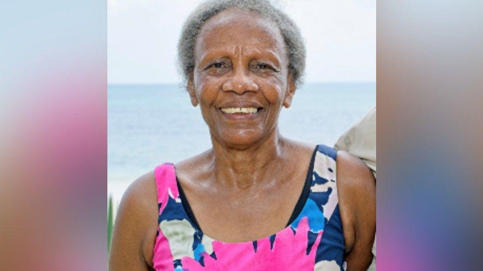 Mel Chevannes photographed by the sea, wearing a pink and blue brightly-coloured sun dress, and looking happy. She has short grey hair and is smiling at the camera.