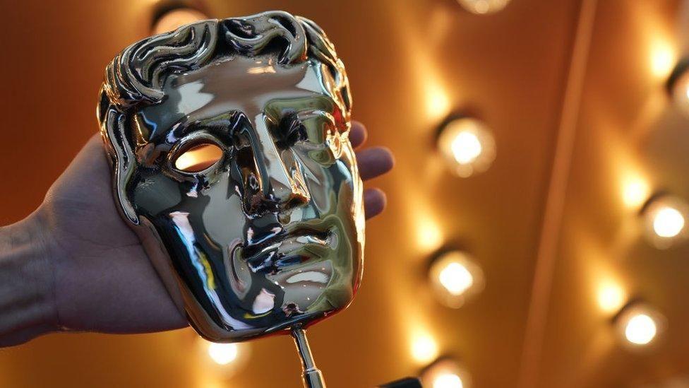 The gold mask trophy used to award BAFTA winners. It is being held up in front of a gold background with LED stage lights.