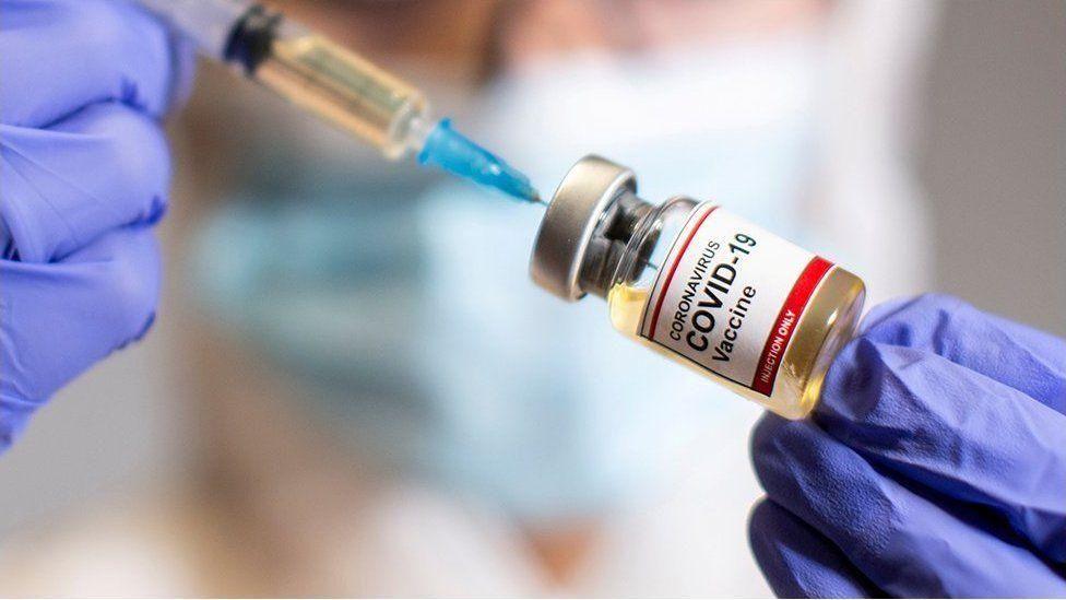 Needle and vaccine in this photo. There's a close up of a dosage in a glass bottle. There's purple gloves holding both sides of the needle and vaccine dose. In the background and out of focus there's a person in a blue medical mask.
