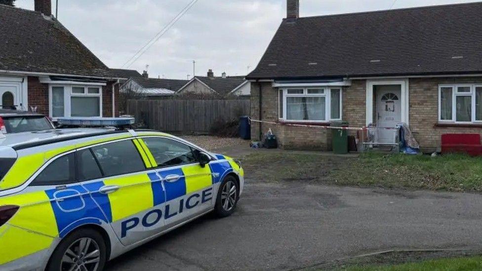 A blue and yellow marked POLICE is parked outside a property. At the front of the property is red and white police tape.