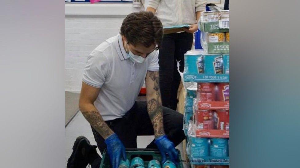 Liam Payne is wearing blue gloves and kneeling on the floor of a room. He is holding a crate full of baked beans. To his right, there are stacks of tinned food and a woman stands behind him