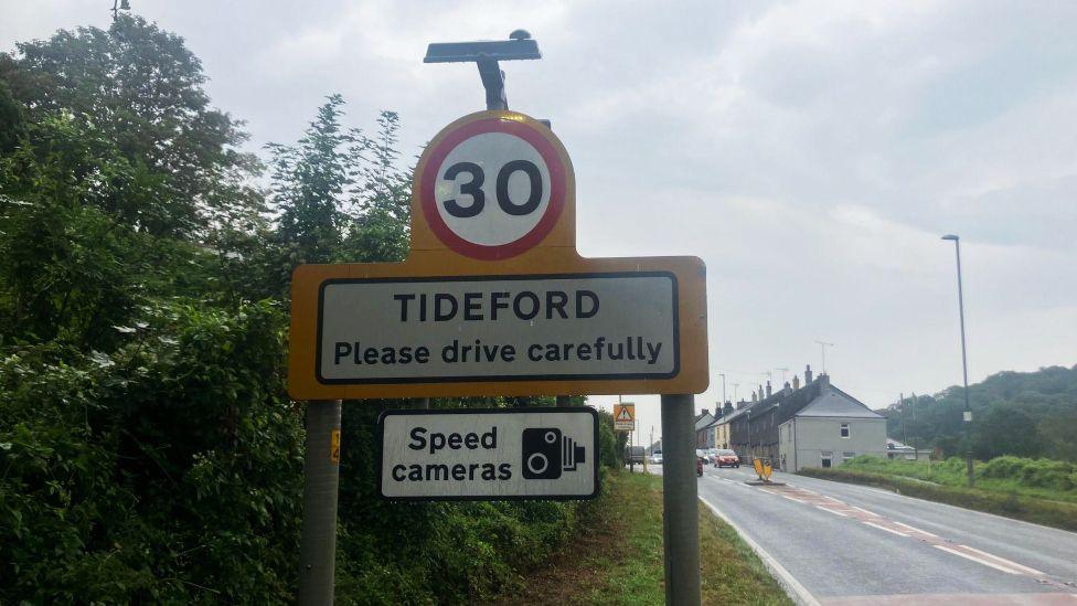 A road speed limit sign which says "30", and, below it 'Tideford, please drive carefully". Below that is a sign saying "speed cameras" and a black and white graphic of a camera. Beside the sign on the right is the road.