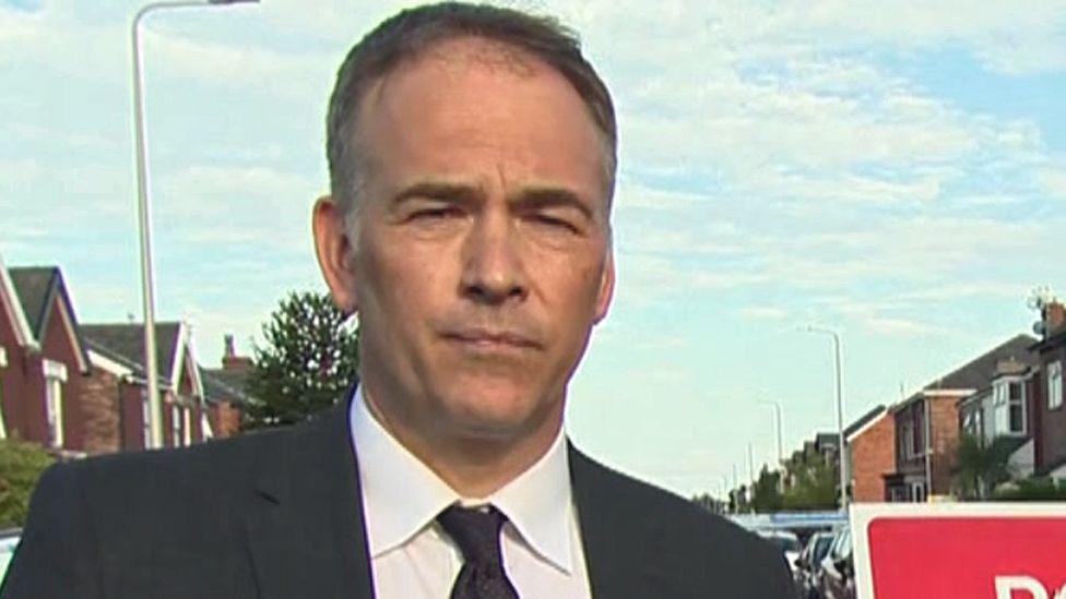 Roger Johnson, in a black suit with a white shirt and dark-coloured tie, stands in a street with a blue sky above