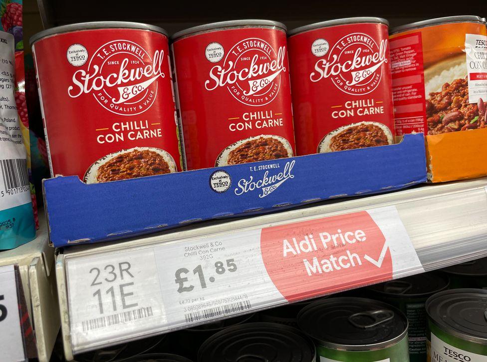 Tins of Chilli con Carne, with a red label reading 'Stockwell and Co' with a picture of mince with rice, on a Tesco supermarket shelf.  On the edge of the shelf is a white label with the price of £1.85 in black writing, and a red logo which says Aldi Price Match with a tick. 