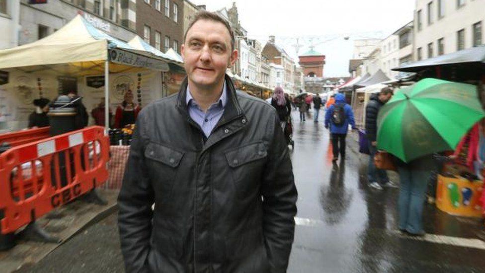 Martin Goss, wearing a black jacket over a shirt while walking down Colchester High Street. It is a rainy day and there are market stalls either side of him, with people walking behind him. He has short brown hair and is looking into the distance with a slight smile.