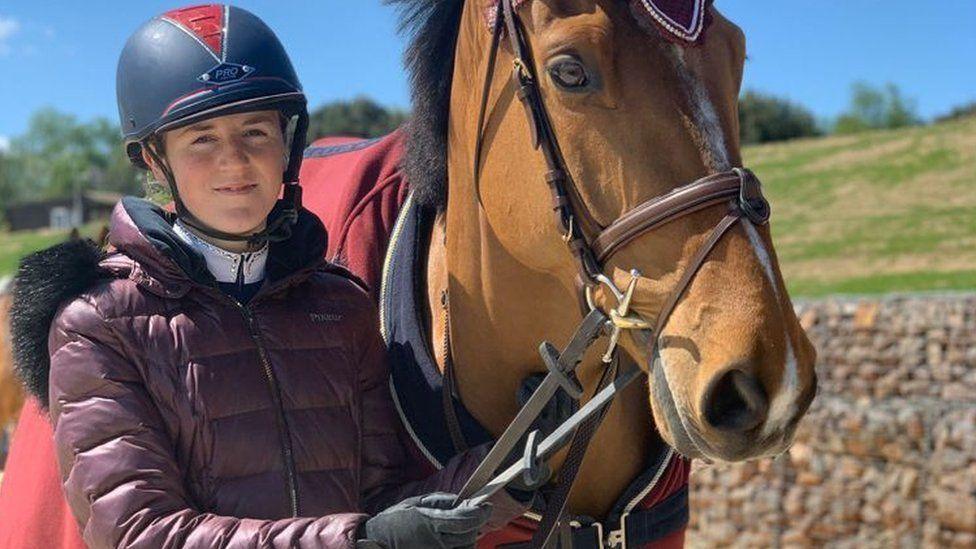 Smiling Brodie, aged 16, stands with a horse, whose reins she is holding. She has a riding helmet on and a plum-coloured coat