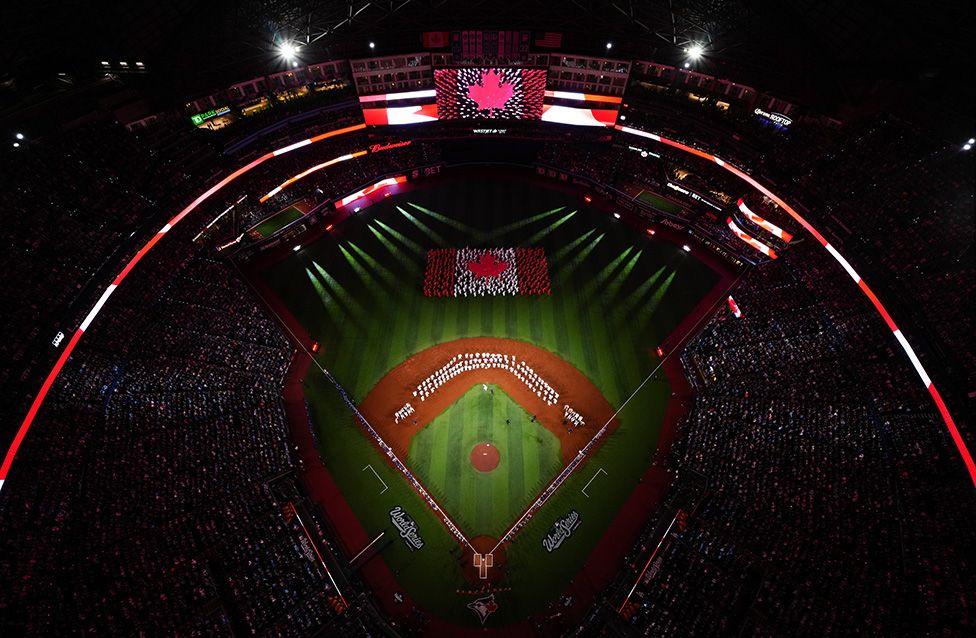 Aerial view of a baseball stadium during a ceremony, with performers forming a Canadian flag on the field under bright spotlights, surrounded by a packed audience and large screens displaying the same flag.