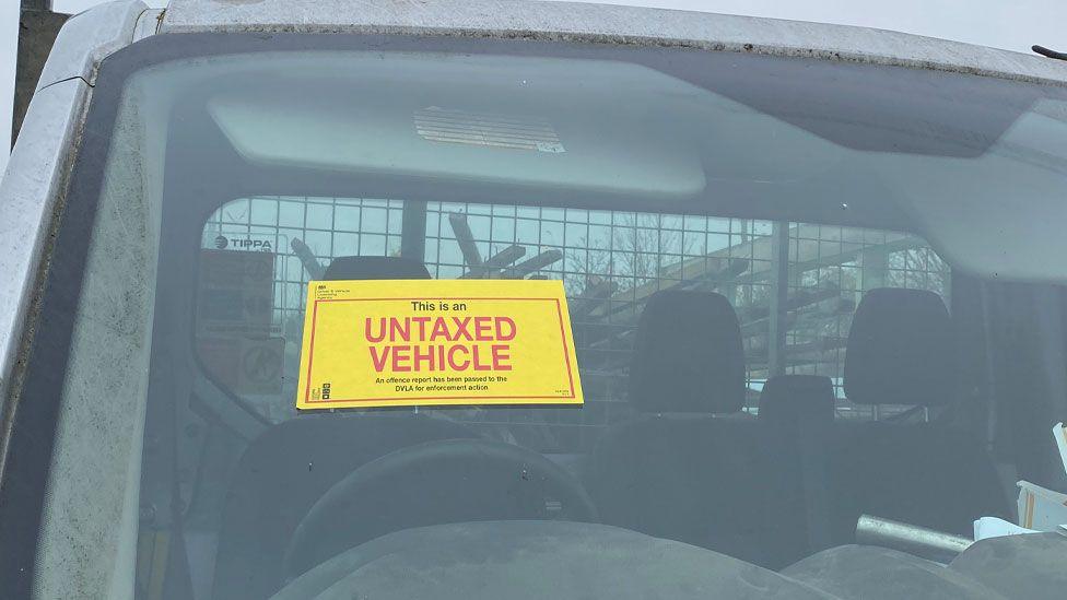 A close up of a vehicle windscreen. It has a bright yellow sticker above its steering wheel. It says This is an untaxed vehicle in black and red lettering