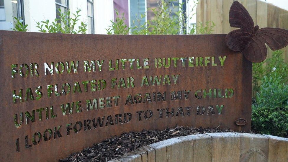 A big bronze-coloured metal plaque with a large metal butterfly on it reads, "For now my little butterfly has floated far away, until we meet again my child, I look forward to that day".