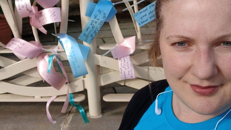 Laura Thompson standing in front of railings. She is on the far right of the image, her hair is back, she is wearing a blue T-shirt and a black top and looking towards the camera. On the cream metal railings to her left are tied blue and pink ribbons, each with writing on them