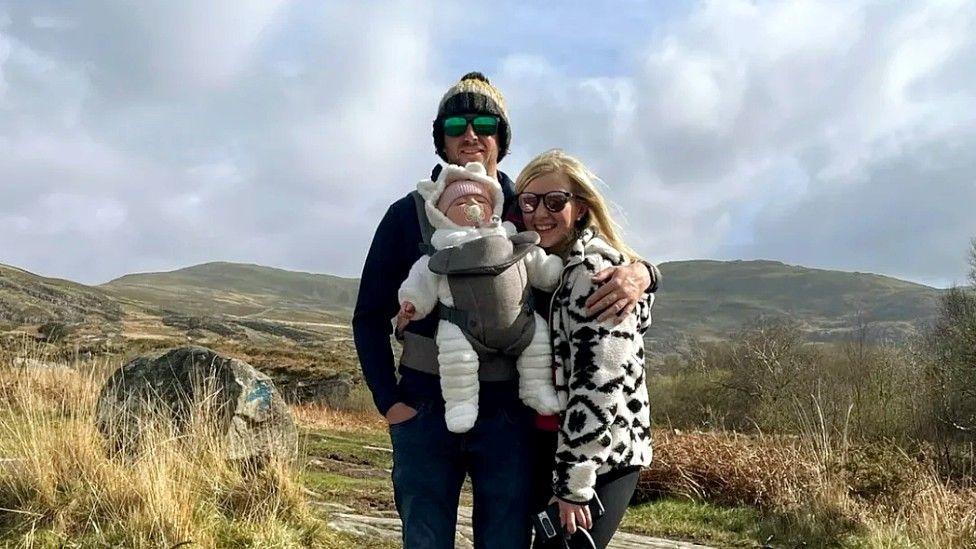 Enlli is standing to the right of her partner, who is carrying baby Cali in a sling. She wears a white fluffy jacket with geometric black pattern on it and sunglasses and black trousers. Her partner has a dark winter jacket and dark trousers, and wears a woolly hat and sunglasses. The baby is wearing a warm all-in-one suit in white, with the hood up and a woollen hat underneath.