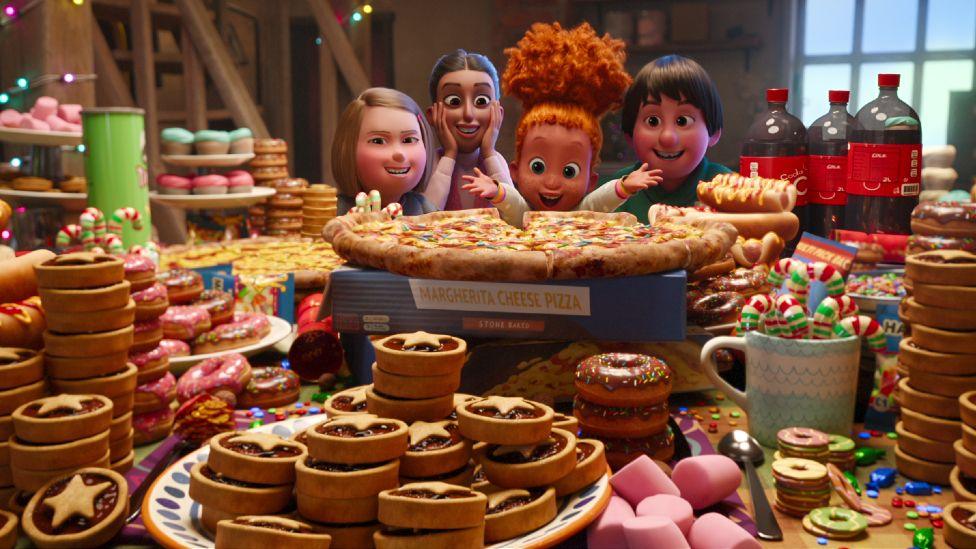 Children looking delighted as they look over a table filled with pizza, fizzy drinks, doughnuts, mince pies and other Christmas food, in a promotional still from Netflix's That Christmas