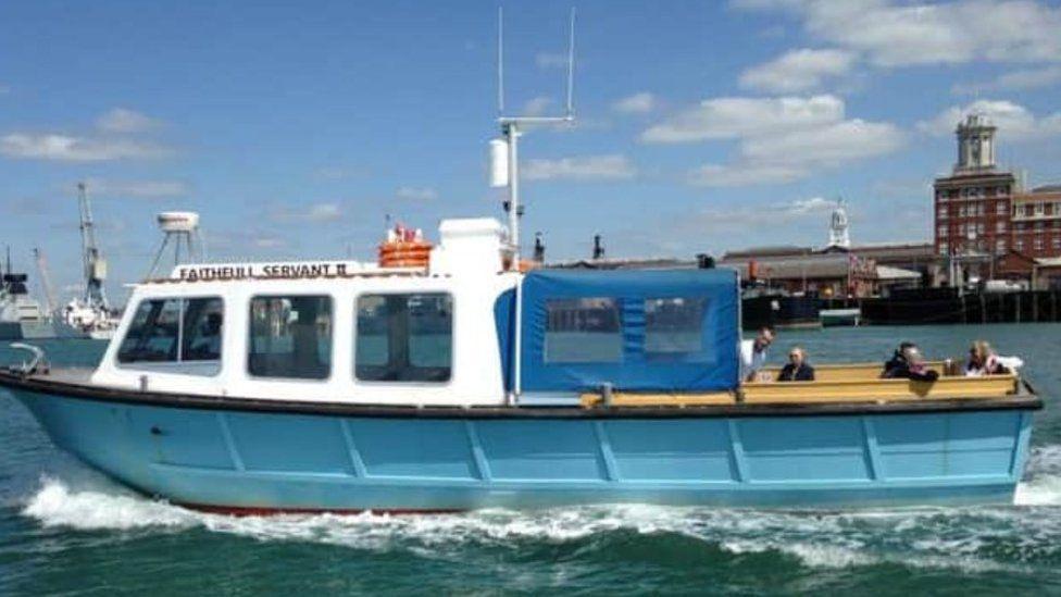 Pale blue and white boat with passengers sat at the rear - it is moving from right to left - buildings and ships can be seen in the background.  It is a bright, sunny day.