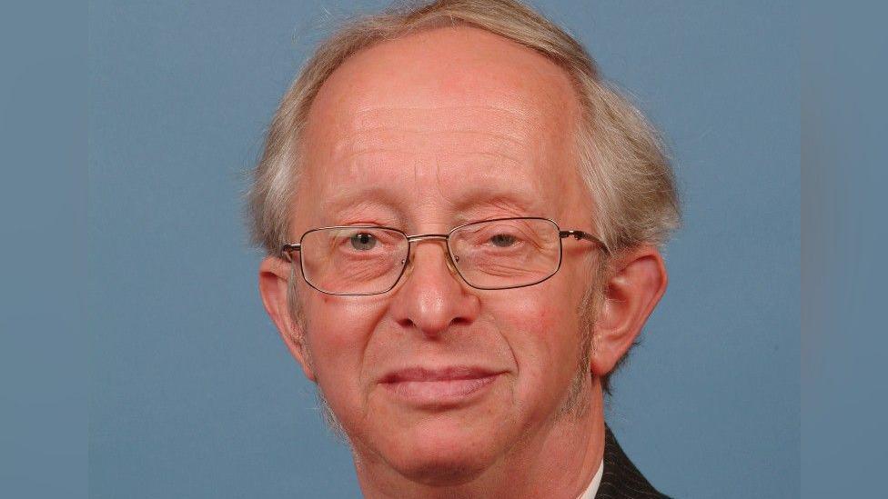 A close-up showing the head of a man wearing silver wire-framed glasses. He has grey hair in a side parting and the collar of a dark suit and white shirt can just be seen. The image is against a blue background.