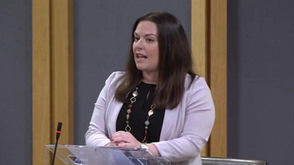 Vikki Howells in the Senedd. She is stood at a lecturn about to speak. She is wearing a black top underneath a lilac jacket and has a beaded and gold necklace on. Her dark shoulder-length hair is untied. 