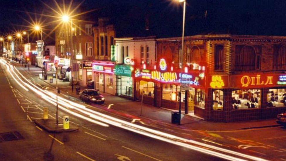 The "curry mile" of restaurants in Manchester - a row of takeaways and restaurants brightly lit at night.