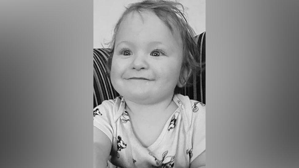 A black and white picture of a smiling Louie Hughes. The baby is wearing a Mickey Mouse vest.