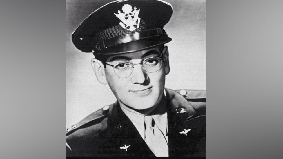 Glenn Miller pictured in black and white, wearing an army uniform with silver buttons on the shoulders. He is also wearing a hat with a large emblem on the front, and a pale shirt and tie. He had metal framed round glasses and is smiling with his mouth closed and head cocked slightly to one side.