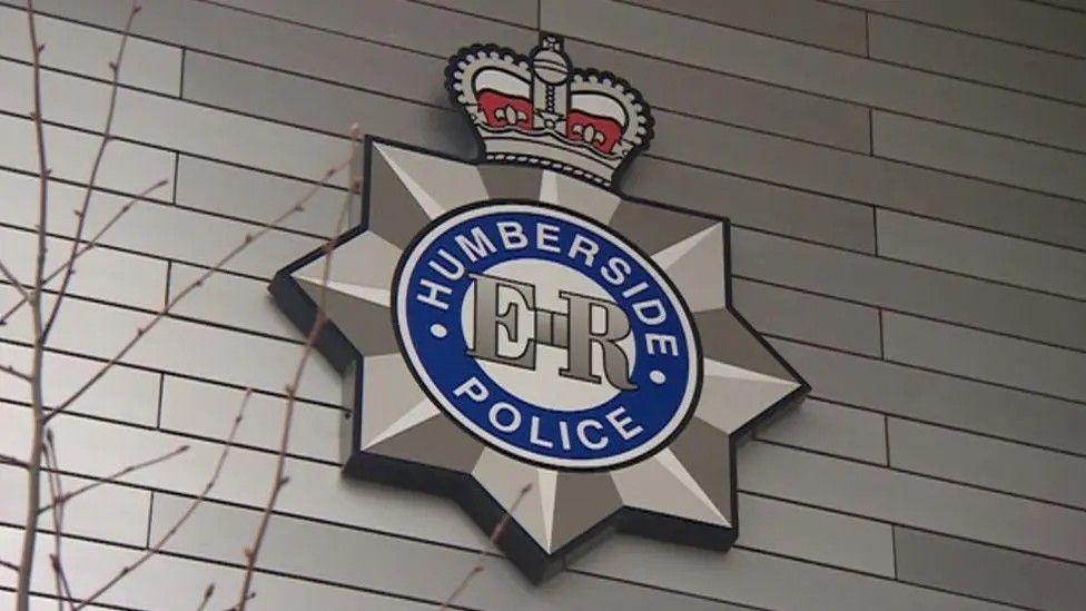 The Humberside Police logo - featuring a blue circle, a silver star and a red crown - on the side of a building. A tree branch is in the foreground of the image and the building is covered in grey cladding