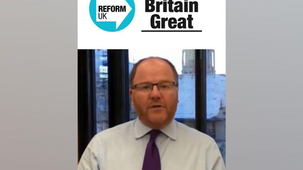 A blurry screengrab showing George Freeman under a Reform UK banner. He is sitting in front of a window, has short greying hair and a reddish close cropped beard. He is wearing glasses, a white shirt and a purple tie.