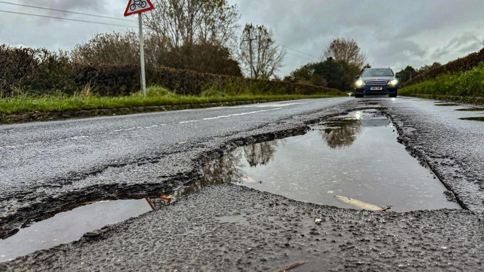 A large pothole on a road. There is water in the pothole and a car driving towards it. There are also trees and bushes on either side of the road. 