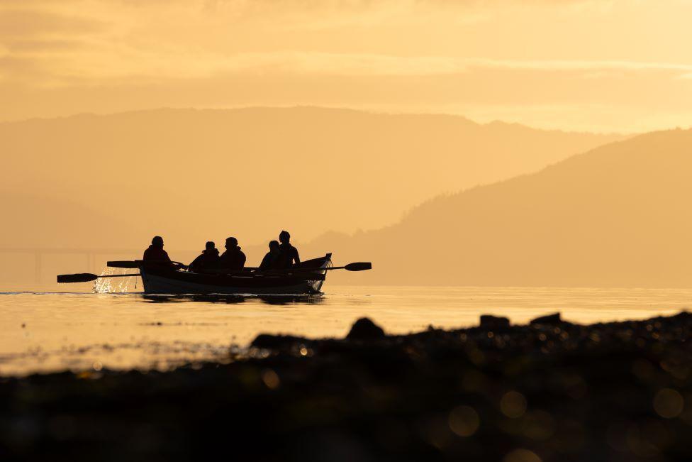 Silhouette of a rowing boat with five people in it, and oars out to the side, with some water splashing, in early evening light, with land of different shades in the foreground and background.