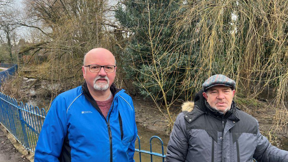 Two men stood in-front of a beck fenced off by a blue metal barrier. The man on the left is wearing a blue jacket, hes wearing glasses, he's bald. The man on the right has a grey flat cap on he's shorter and is wearing a grey jacket. 

