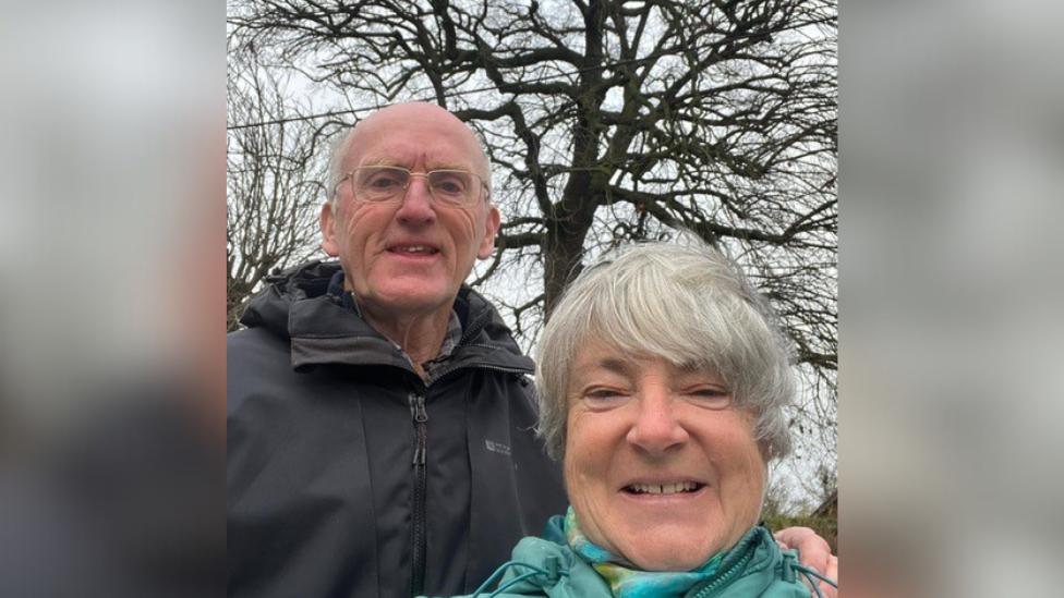 Philip George wearing a black coat and glasses in front of the large oak tree. He is stood next to his wife who has grey hair and is smiling while wearing a blue coat and scarf.