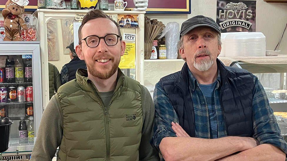 Matt and Barry Howarth standing in the shop. Matt has short dark hair and a beard and black framed glasses and is smiling. His dad has a white goatee and is wearing a baseball hat