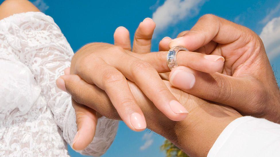A close-up of a groom putting a wedding ring on a bride's finger - blue sky with a few clouds can be seen behind them.