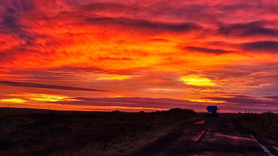 Red and orange sunset sky, with a car in the distance, its red tail lights visible.