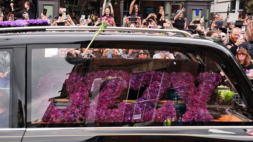 A picture of the back of the hearse showing an "Ozzy" floral tribute alongside the coffin, and crowds behind it