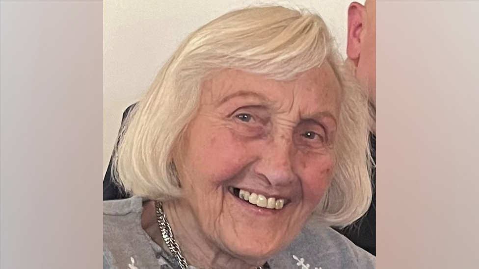A family photo of Elizabeth. She has brown eyes and shoulder length white hair. She is smiling at the camera. She is wearing a grey cardigan and a silver necklace.