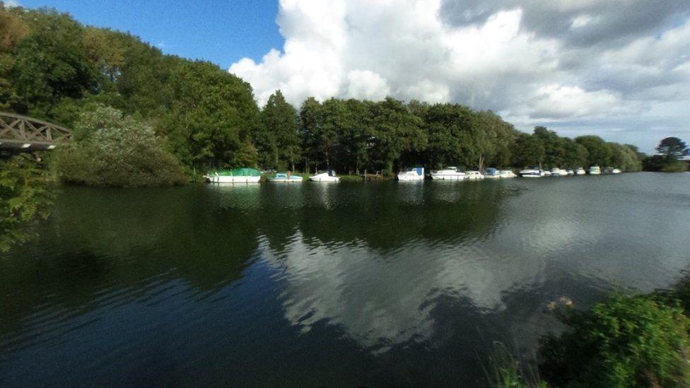 River bank with shrubs bridge over the water to left and boats along a far side bank lined with trees