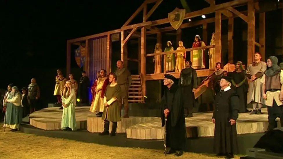 A view of a wooden stage set with at least 20 performers on stage at the end of a Shakespeare play, standing looking towards the audience