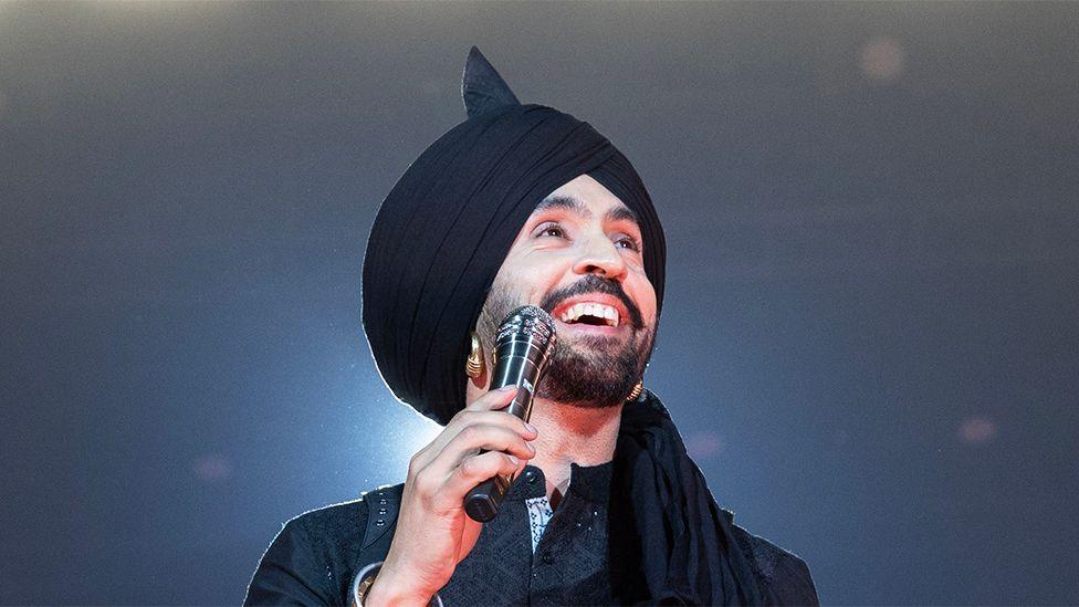 A picture of singer Diljit Dosanjh on stage. He is holding a microphone in his hand while looking up, dressed in all black. Behind him is a spotlight sparkling.