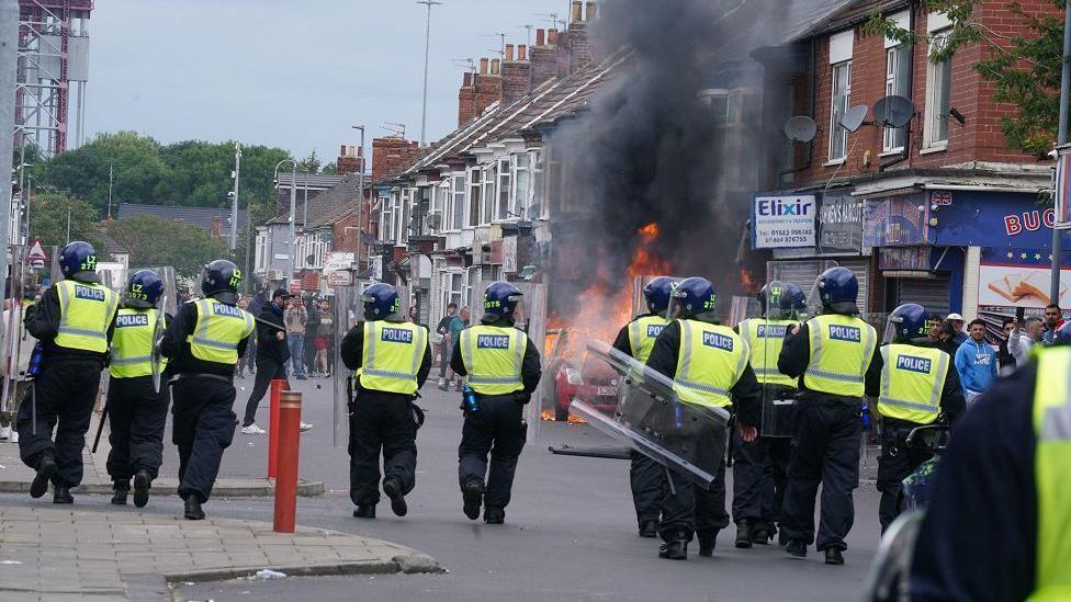 A line of police officers in riot gear approaching a group of rioters. Between the two groups a car is burning in the middle of the high street.