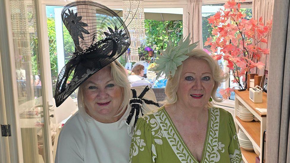 Paula Gamester and a friend standing in her conservatory in the sunshine. both are blonde and Paula is wearing a cream dress with a large wired black hat with flowers in the pattern. Her friend is wearing a sage green dress with white embroidery and a floral fascinator