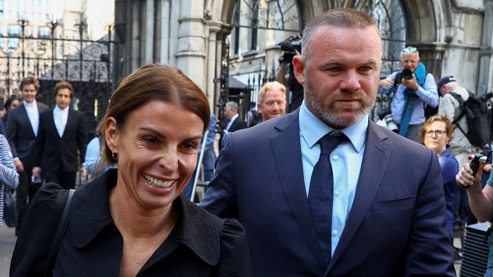 Coleen Rooney smiling next to Wayne Rooney, both walking away from court with photographers in the background on 17 May 2022