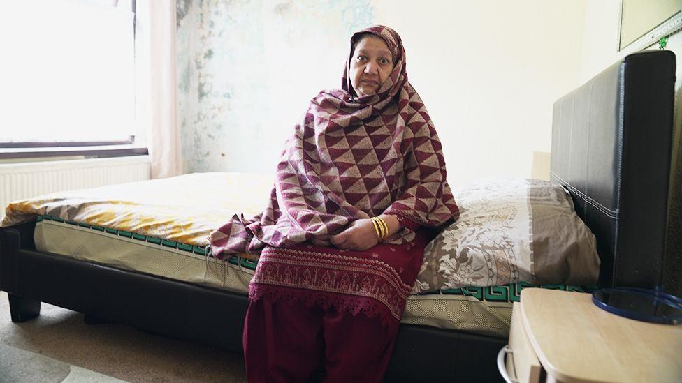 Bushra is sitting on the edge of a bed in a modest room. The bed has a dark frame with a mattress covered by patterned bedding in shades of yellow, gray, and green. Bushra is wearing a maroon outfit with a shawl featuring a red and beige geometric triangle pattern and gold bangles on the wrist. A small wooden bedside table with a blue item on top is visible to the right, and the wall behind the bed shows signs of peeling paint and discoloration.