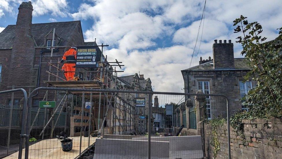 Council workers installing a covered walkway on the bridge over the River Cocker. A metal fence has been placed across the footpath while the work is being carried out. The walkway is made of scaffolding.