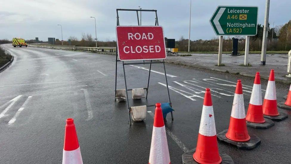 A line of cones and a road closed sign blocking the entrance to a section of dual carriageway, with a police van parked further along and a road sign indicating the route heads towards Leicester and Nottingham