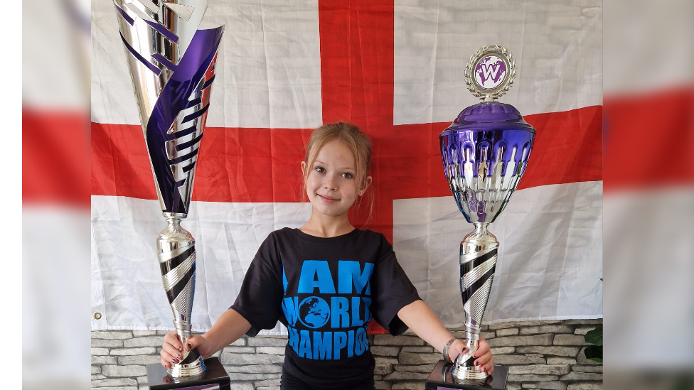 Isla, a 12-year-old dancer, has her blonde hair back in a ponytail and is wearing a black t-shirt with 'I am a world champion' written on it. She is holding two bit trophies, which are silver, black and purple in colour. She is standing in front of an England flag and smiling at the camera.
