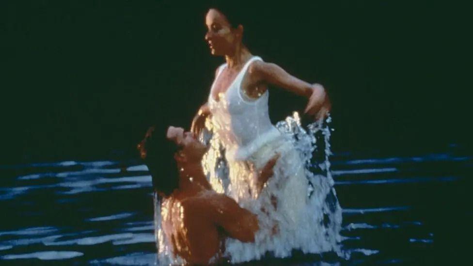 A still shot from the film Dirty Dancing. Patrick Swayze is seen holding the waist of actor Jennifer Grey as he lifts her up out of the water they are both in. He is topless and she is wearing a white all in one swimsuit